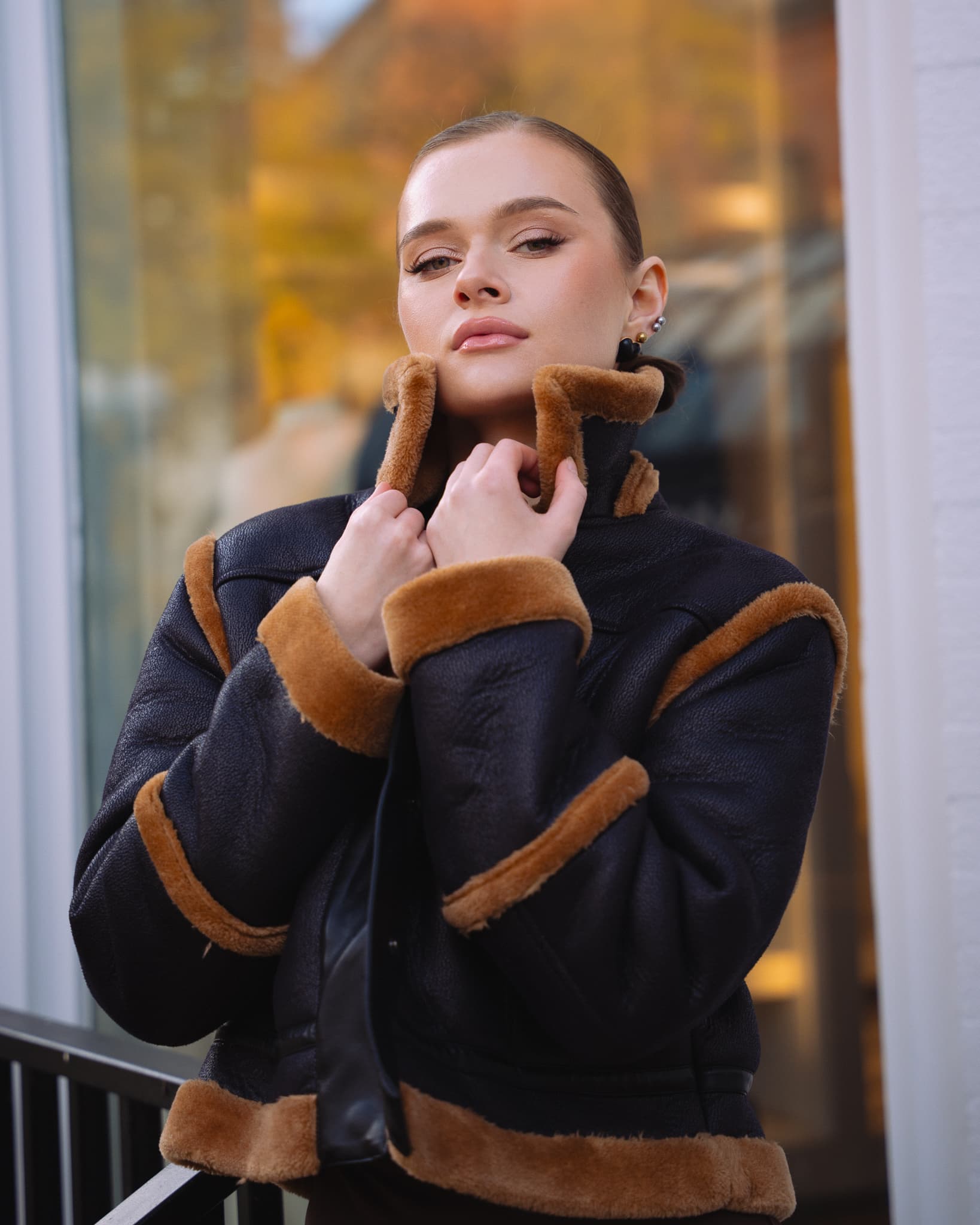 Woman in a shearling jacket touching her collar by a window